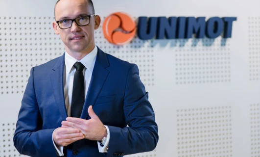 Pracownik umysłowy w formalnym stroju składającym się z marynarki i koszuli z logo UNIMOT. Profesjonalny strój do pracy w branży technologicznej.